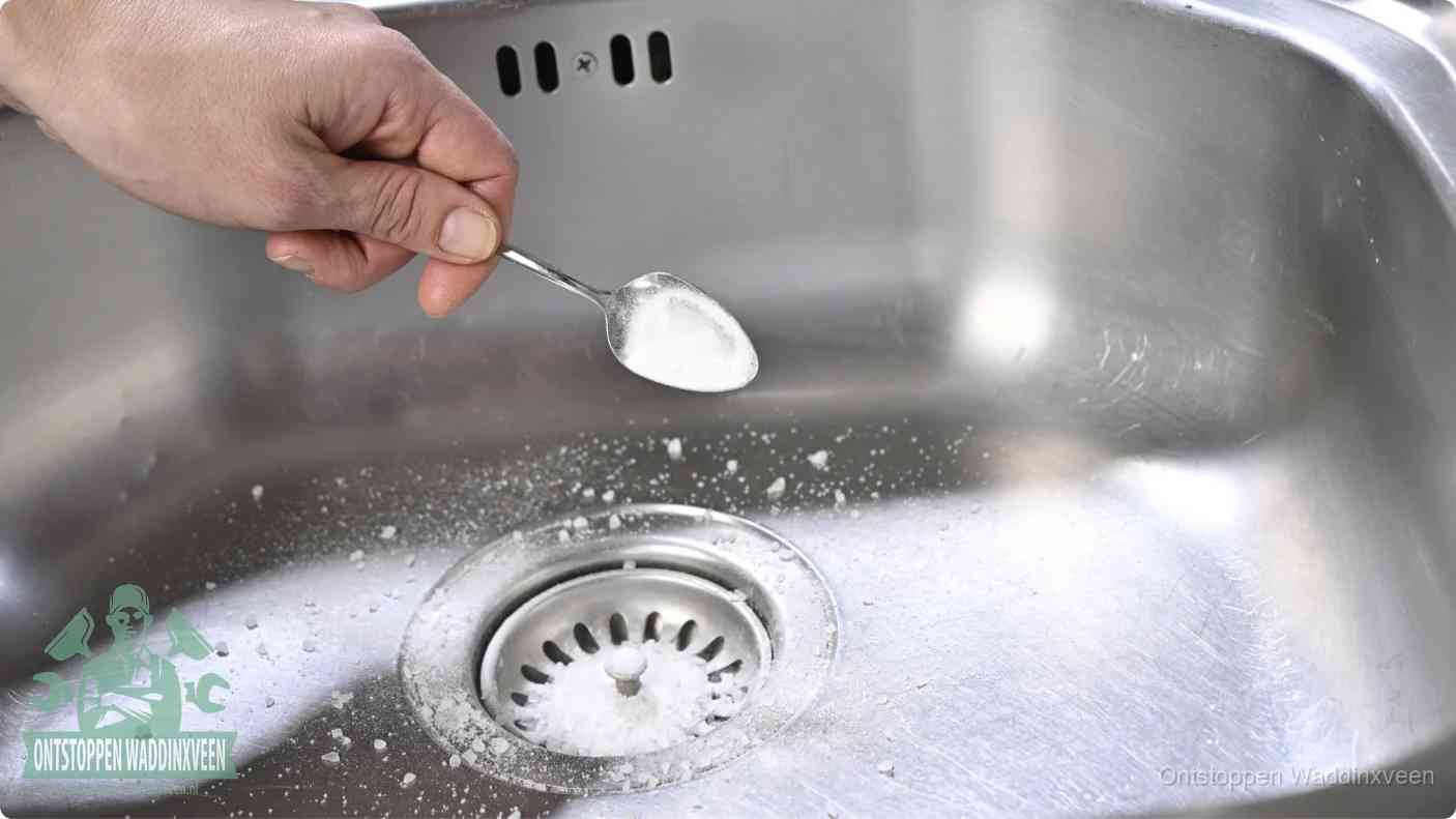 Een hand strooit met een lepel soda in de afvoer van een roestvrijstalen spoelbak, de soda en azijn huismiddel-methode om de afvoer te ontstoppen in Waddinxveen