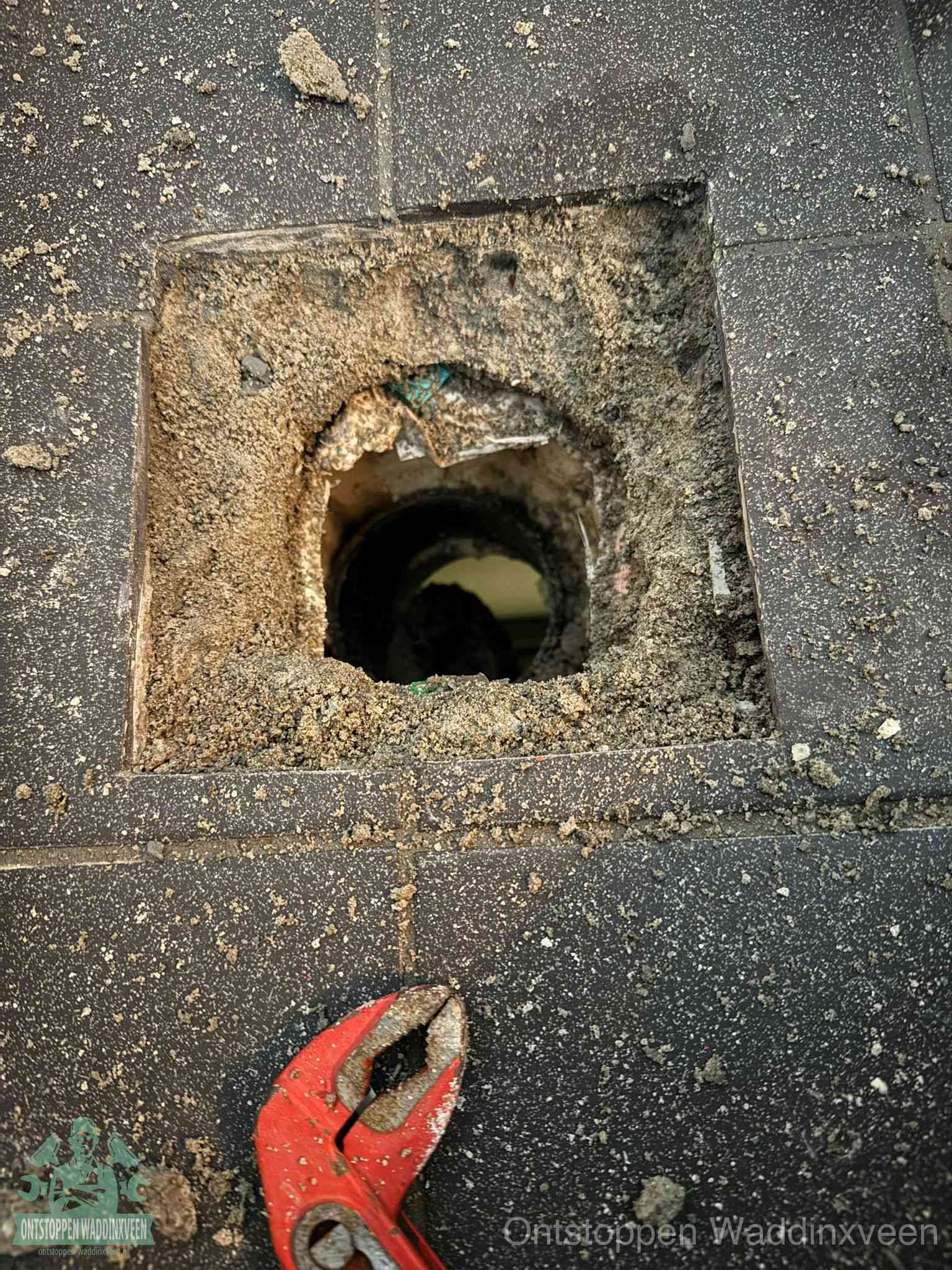 Een geopend rioolputje in een betontegelvloer met losgemaakt rooster en een zichtbare ronde afvoerbuis, het achterhalen waar de afvoerverstopping zit in Waddinxveen