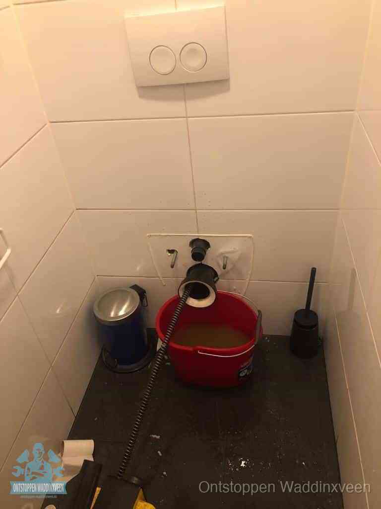 Een ontstopte wand-wc met verwijderde toiletpot waar een ontstoppingsveer in de wanduitgang verdwijnt, het achterhalen waar de verstopping zit in Waddinxveen