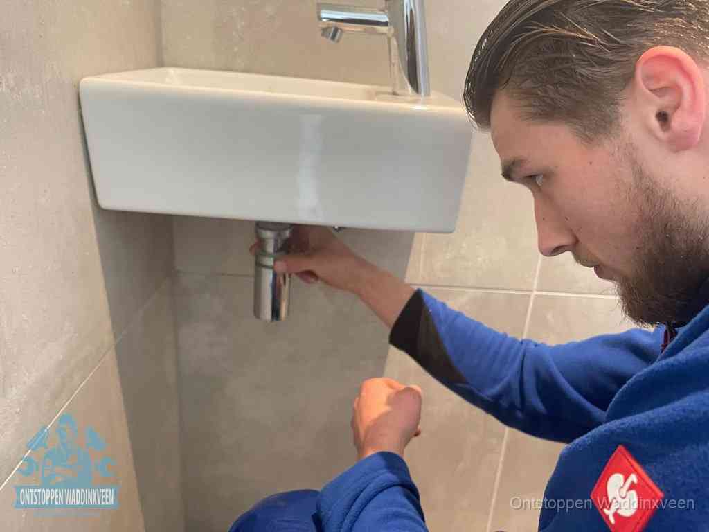Een monteur draait met de hand de zwanenhalssifon onder een fonteinwasbak in de toiletruimte los om de bron van rioollucht in Waddinxveen te vinden