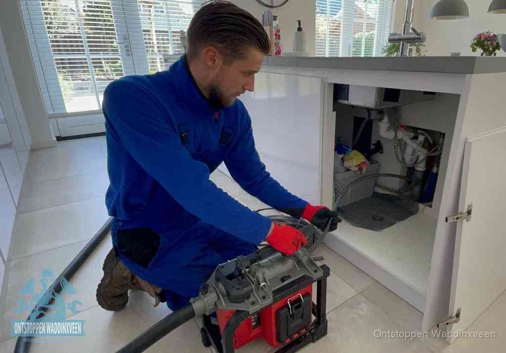 Een monteur voert een professionele elektrische ontstoppingsmachine in de afvoer onder de spoelbak tijdens het gootsteen ontstoppen in Waddinxveen